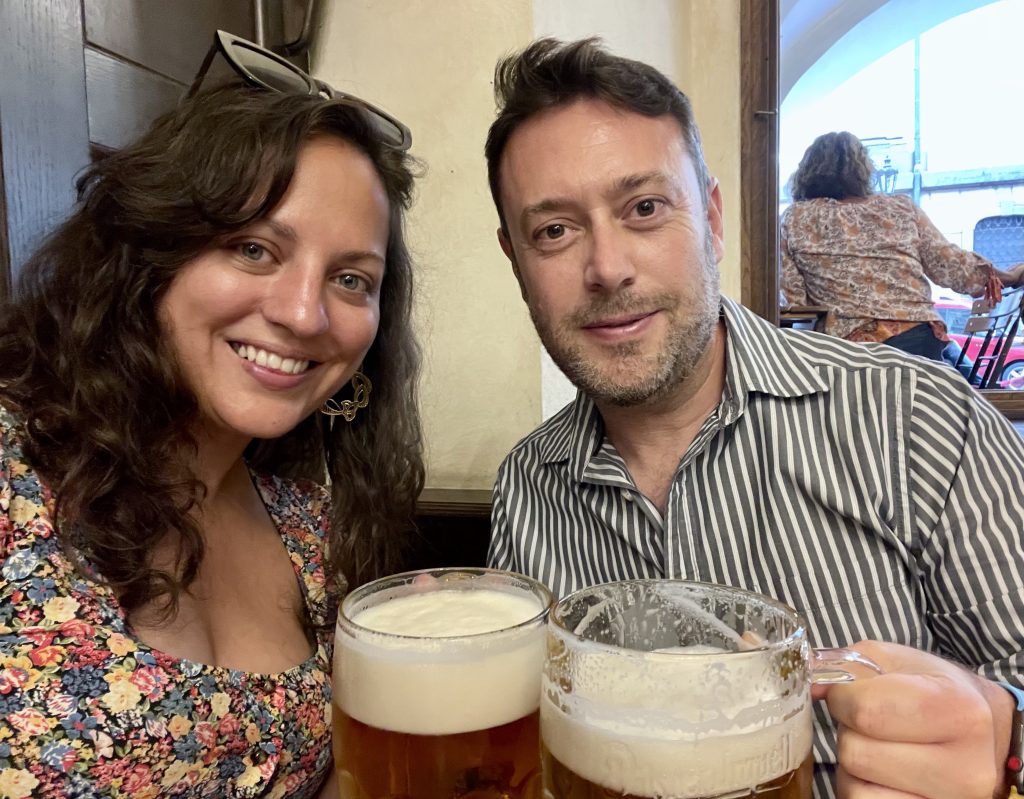 Kate and Charlie hold us two foamy Czech beers and smile. Kate has long curly hair and wears a flowered dress with a square neckline. Charlie wears a gray and white striped long-sleeved shirt.