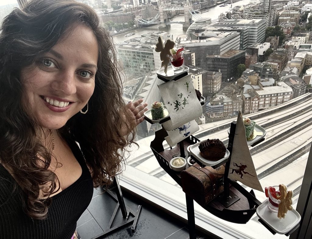 Kate smiles in front of a small pirate ship topped with afternoon tea cakes.  Behind her is the London skyline, including Tower Bridge.