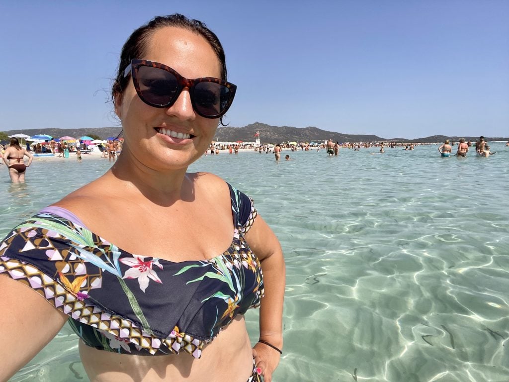 Kate is taking a selfie, standing in shallow bright clear aquamarine water, wearing a navy blue bikini top covers with tropical flowers.