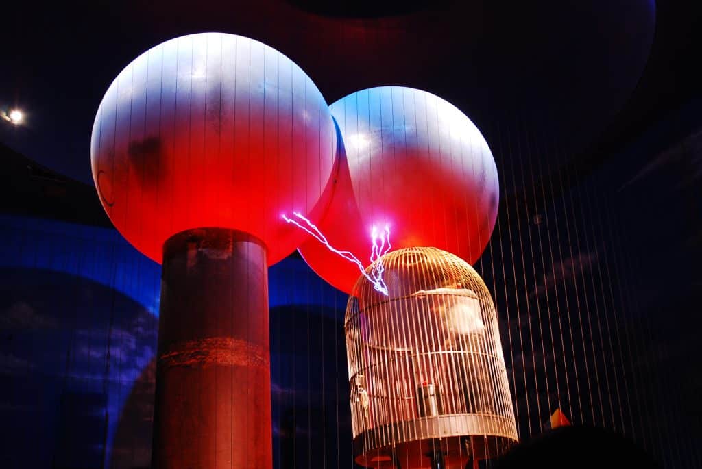 Two bright red glowing orbs in a dark room sending what looks like a strike of lightning to a giant bird cage with a man inside it.