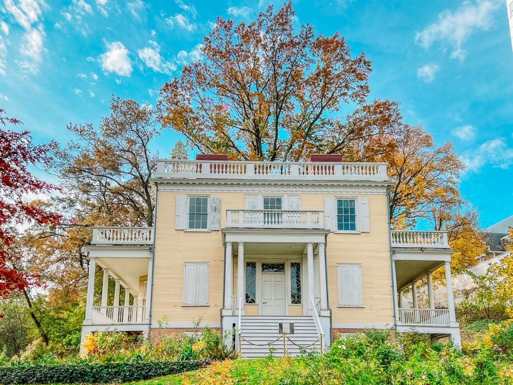 Yellow house with white shutters and a fall tree and blue sky behind it