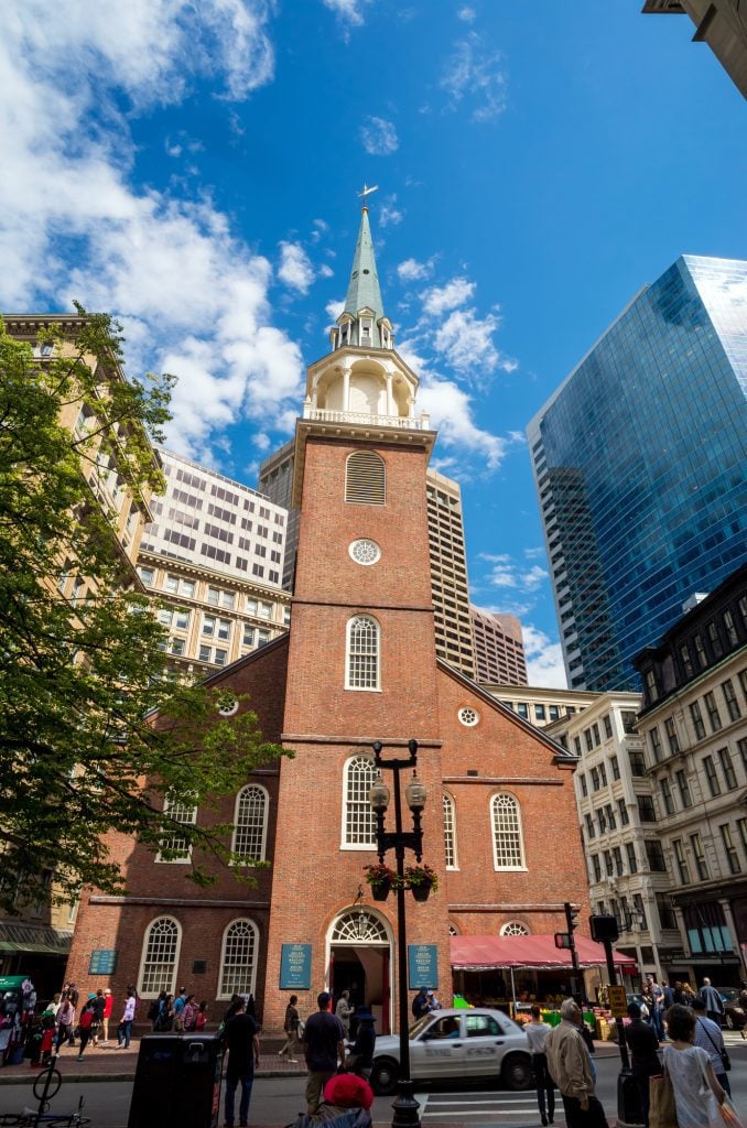 An old-fashioned brick church with a white bell tower surrounded by modern skyscrapers.