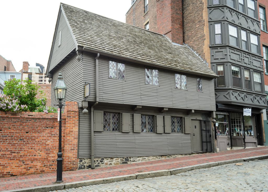 A small gray house from the 1700s with tiny windows, set amongst taller brick buildings on a cobblestone street.