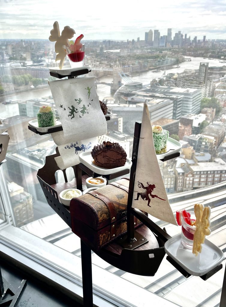 A model of a pirate ship. On it are balanced several pastries, including a fairy perched on a red pudding with an alligator gummy coming out of it. In the background you see a view of gray London from above, including the Tower Bridge.