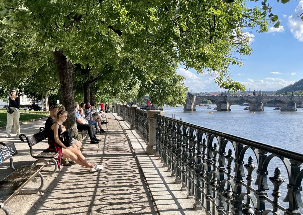 People sitting on benches underneath leafy trees, facing the Vltava River in Prague.