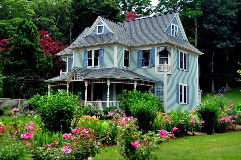 A Victorian house with a wraparound porch, painted shades of green, yellow, and blue. There is a well-manicured flower garden in front.
