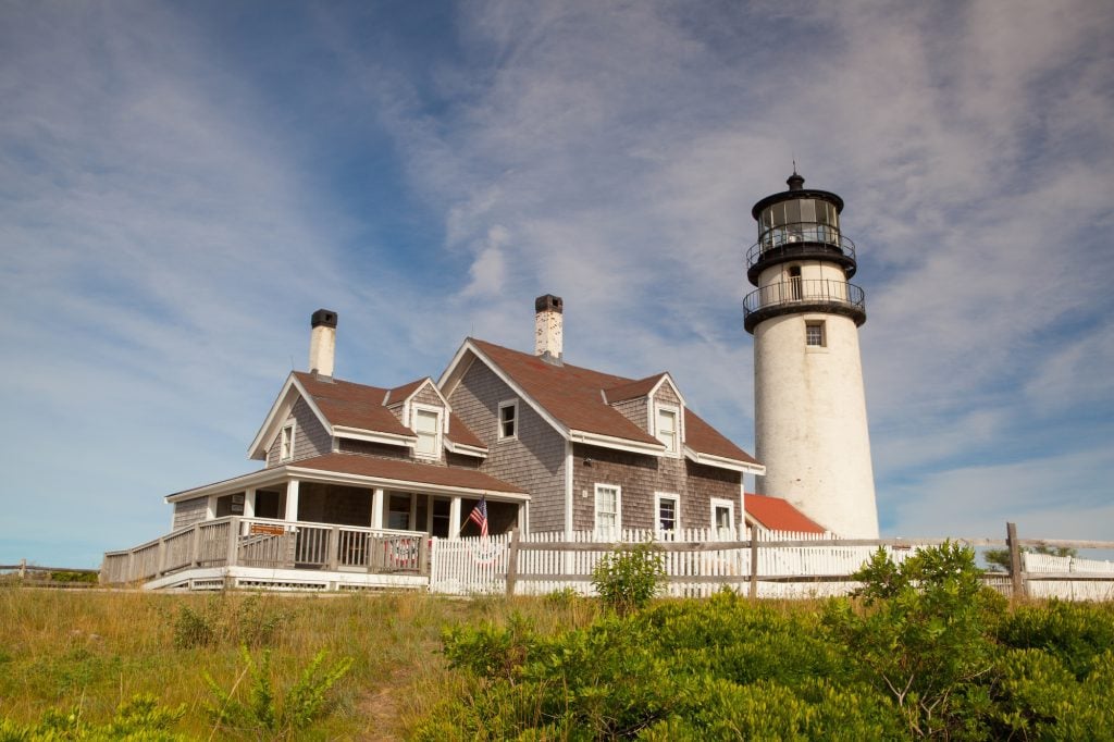 A small gray cottage with a white picket fence next to a white lighthouse.