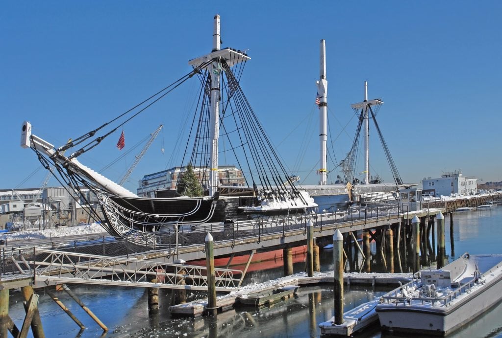 An 18th century old-fashioned large black ship docked in a harbor on a winter day. There is a bit of snow on the ground and a bright blue cloudless sky.