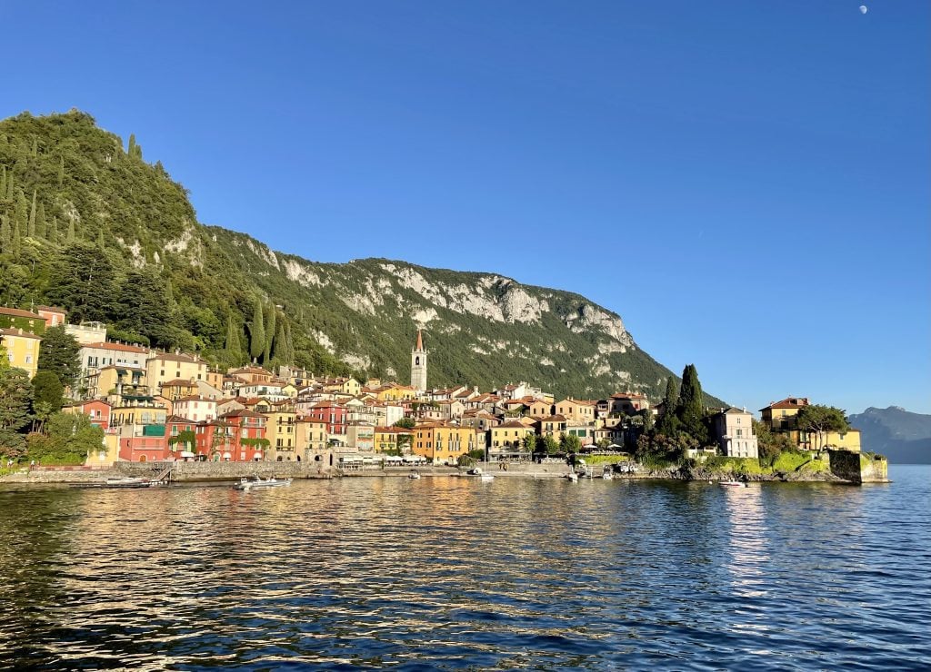 A brightly colored small town with a church tower set on the edge of a massive lake, mountains behind it.