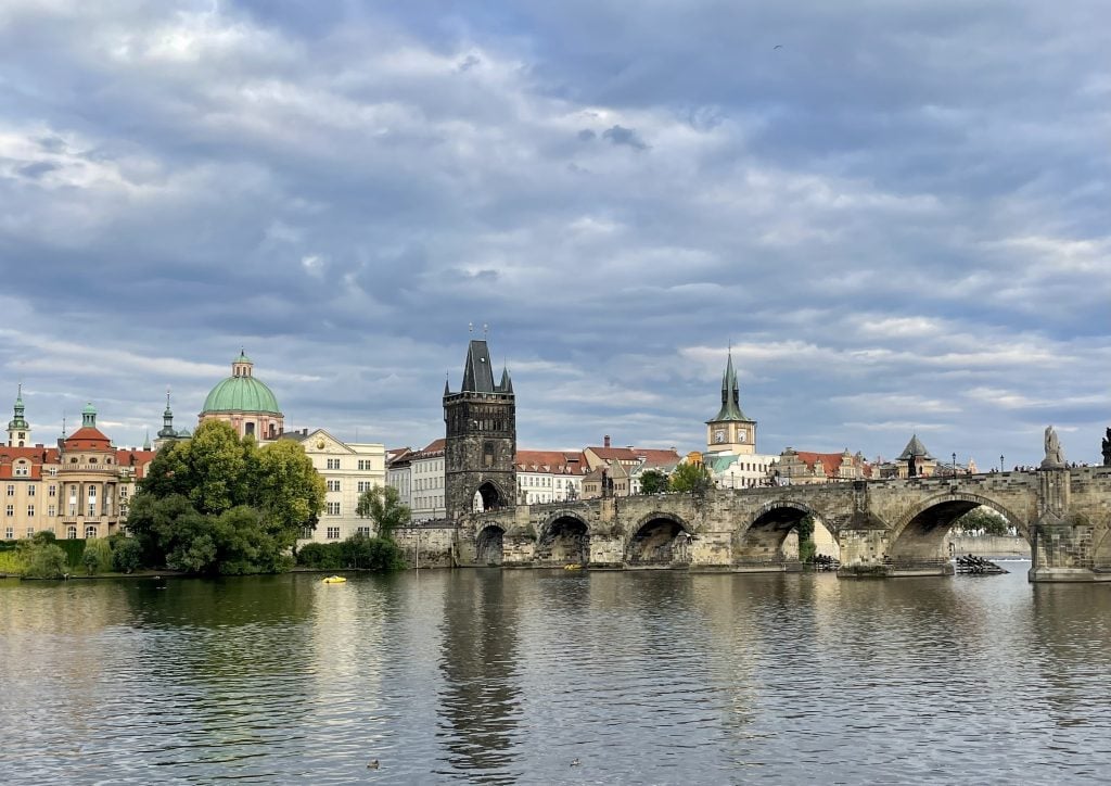 A view of the stone Charles Bridge and several Baroque buildings topped with orange and green roofs in Prague, underneath a cloudy sky.
