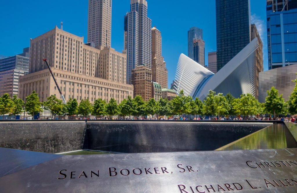 An up close view of the edge of the 9/11 Memorial fountain, with names carved into the edge including Sean Booker, Sr. In the background are the white wings of the Oculus buildings and several modern skyscrapers.