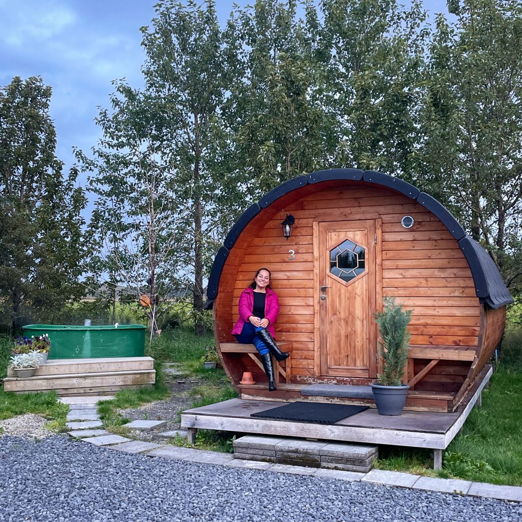 Kate smiles and sits on a bench in front of a small round wooden house, the kind a hobbit would live in. Next to her is a big green hot tub.