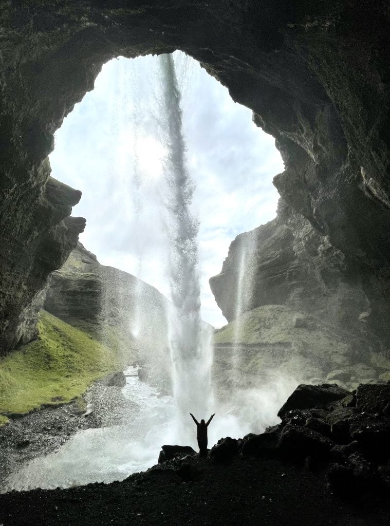 Kate standing silhouetted behind the waterfall, holding her arms up in joy.