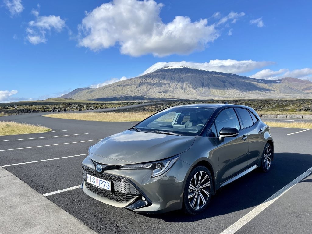 A green-gray Toyota sedan parked alone in a parking lot. Behind it is a mountain topped with a glacier.