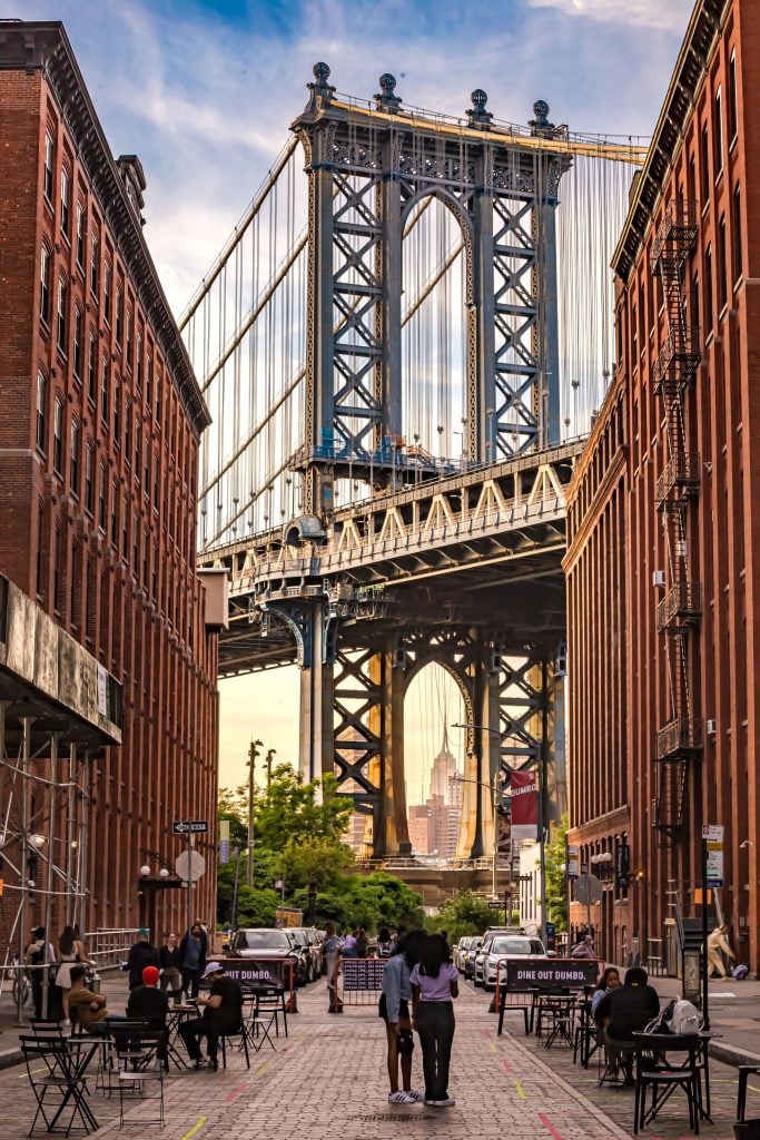 The Manhattan bridge as photographed between two brick buildings, the Empire State Building seen in the distance through the bottom of the bridge.