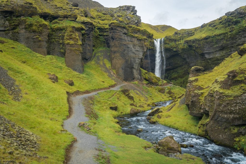 A winding gravel path next to a river in a green canyon leading to a waterfall in the distance.