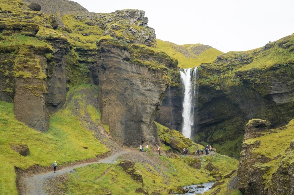 Hikers walking on a gravel path in a green canyon toward a softly falling waterfall.