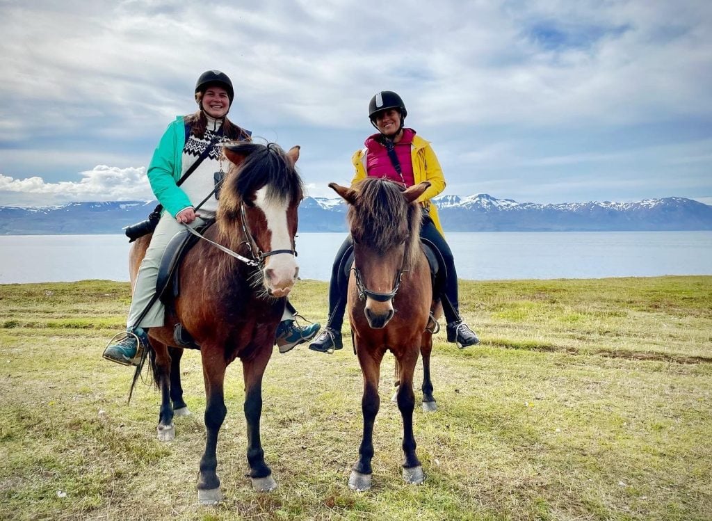 Amanda and Kate on horseback, side by side. The horses are short and brown with shaggy hair, and behind them is a calm bay and snow-covered mountains.