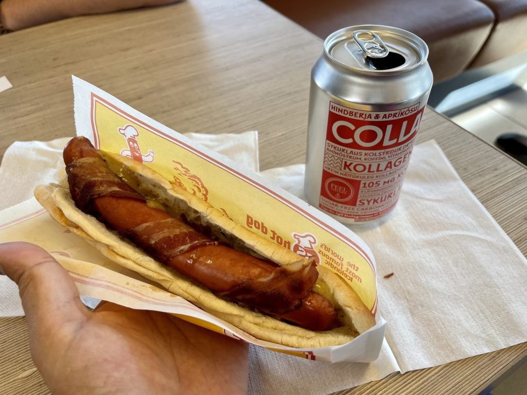 Kate's hand holes a hot dog wrapped in bacon. Next to it is a can of Collab soda.
