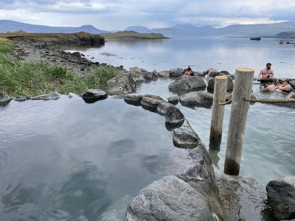 Several natural hot pools next to a misty gray fjord, several people sitting in the pools.