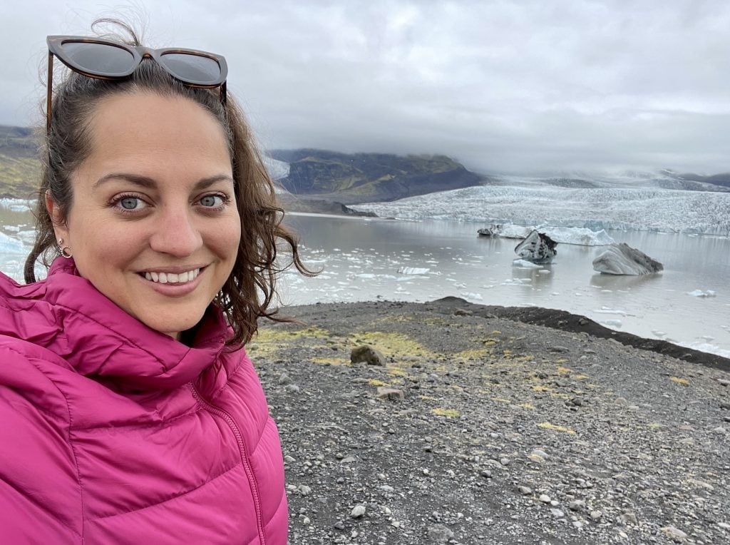 Kate wearing a pink coat and taking a smiling selfie in front of a misty lagoon filled with black-streaked icebergs, a large pale blue glacier on the mountain in the background.