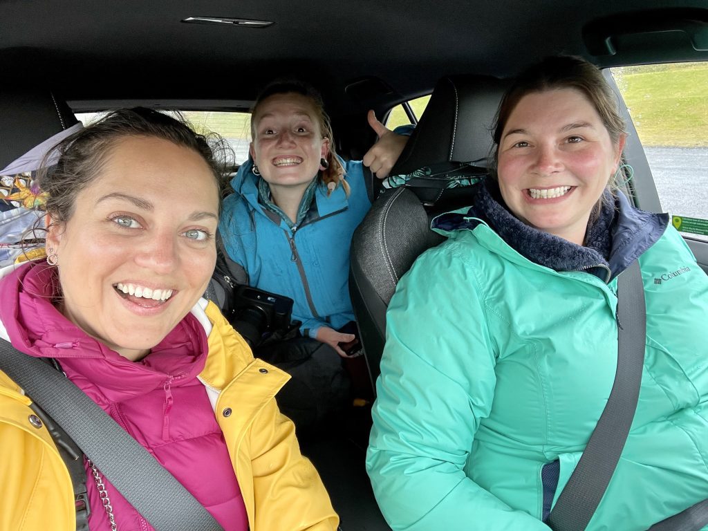 An in-car selfie of Kate and Amanda in the front seats, and a girl in the backseat. All three are wearing brightly colored raincoats and grinning.