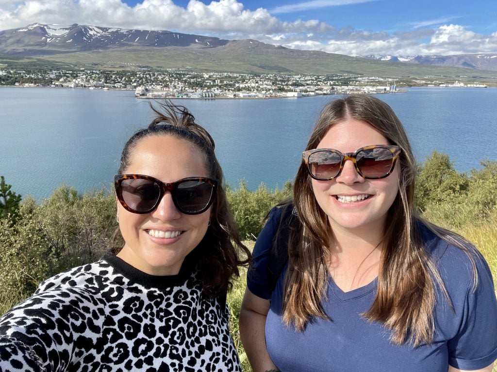 Kate and Amanda taking a smiling selfie, both in tortoiseshell sunglasses. Behind them is a calm blue fjord and the small city of Akureyri, with a mountain behind it and a bright blue sky.
