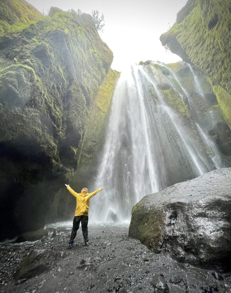Kate stands next to a waterfall wearing a yellow raincoat, black rain pants, and black boots. She holds her arms up in joy and it's clear she's getting very wet.