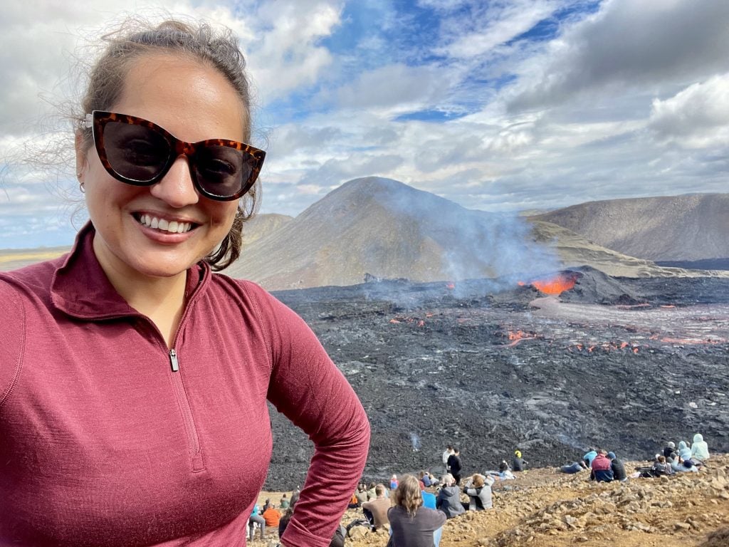 Kate taking a smiling selfie in sunglasses in front of an active volcano spewing bright red lava.