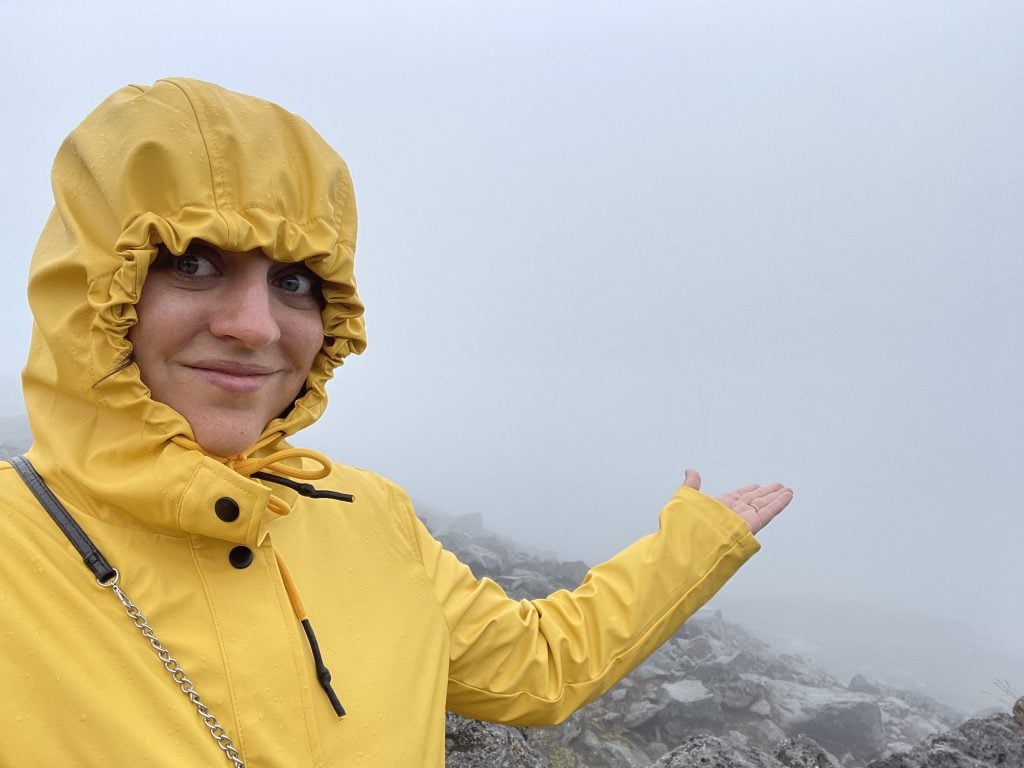 Kate standing in a hooded yellow raincoat, holding her hand out toward a foggy landscape where you can see almost nothing behind her.