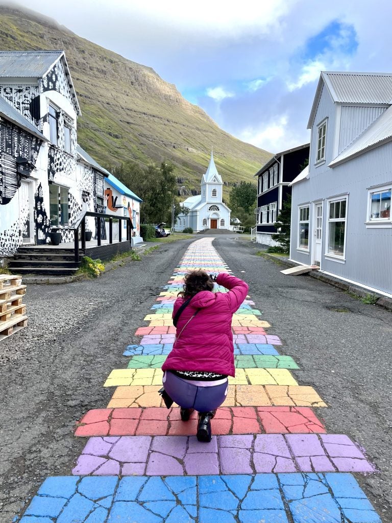 Kate squatting and taking a photo of a rainbow-striped street leading up to a cute pale blue church against a mountain.