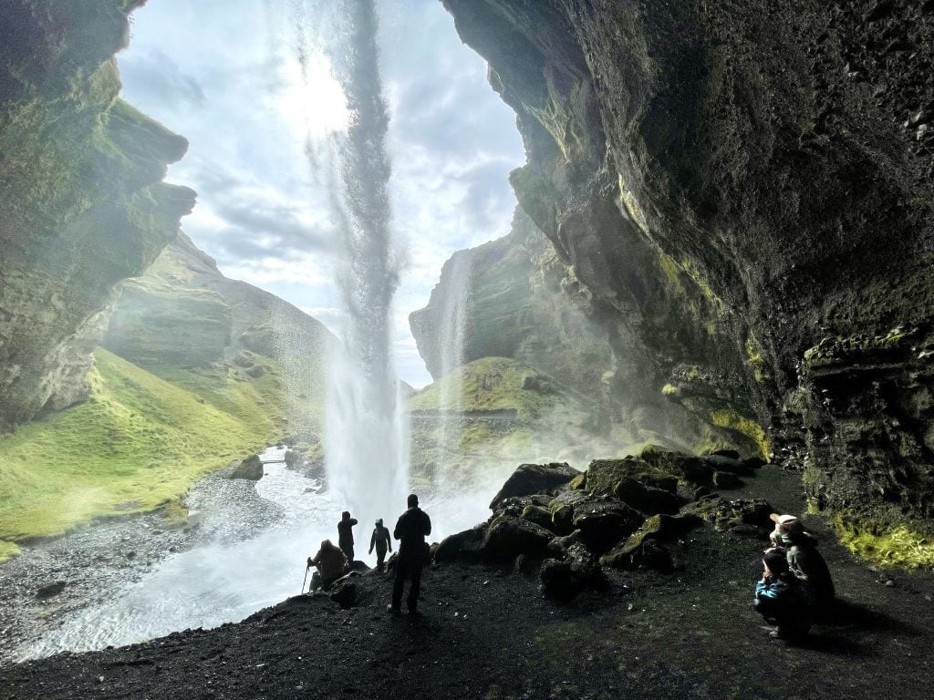 Several people behind a waterfall, taking photos of the mist. Behind the waterfall looks like a large gray cave.