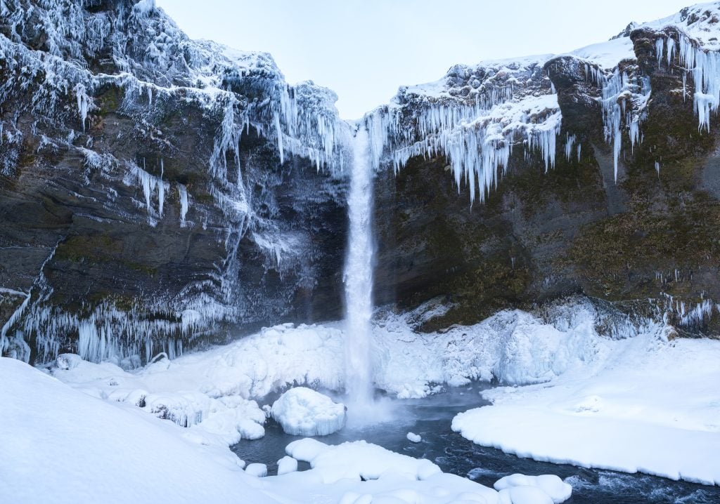 Kvernufoss waterfall surrounded by snow, and the top edges of the cave are covered in hundreds of jagged icicles.