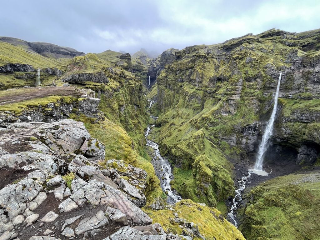 A craggy stone canyon covered with tons of green moss. A river runs through it and there are three skinny waterfalls. Clouds hang in the air.