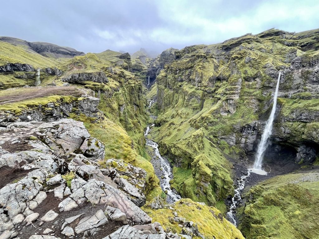 A spectacular craggy canyon covered with green moss, three very tall and skinny waterfalls falling into a river carving through it.