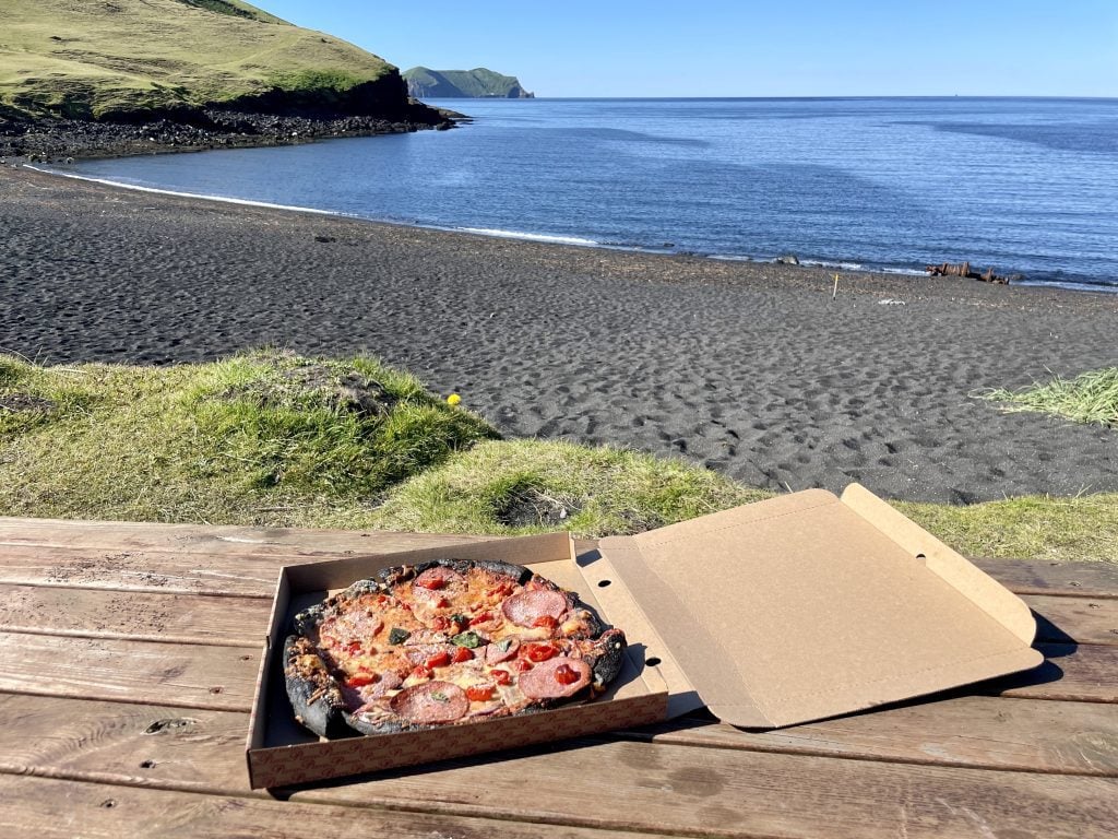 A picnic table with a black crust pizza in a box on it, in front of a black sand beach next to a calm bright blue sea.