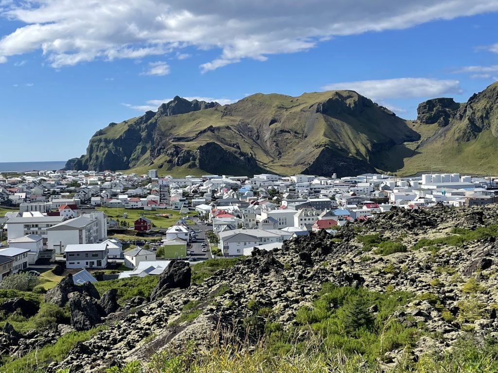 A small city with hundreds of buildings. In the background is a craggy green mountain and in the foreground is rough gray dried lava interspersed with greenery.