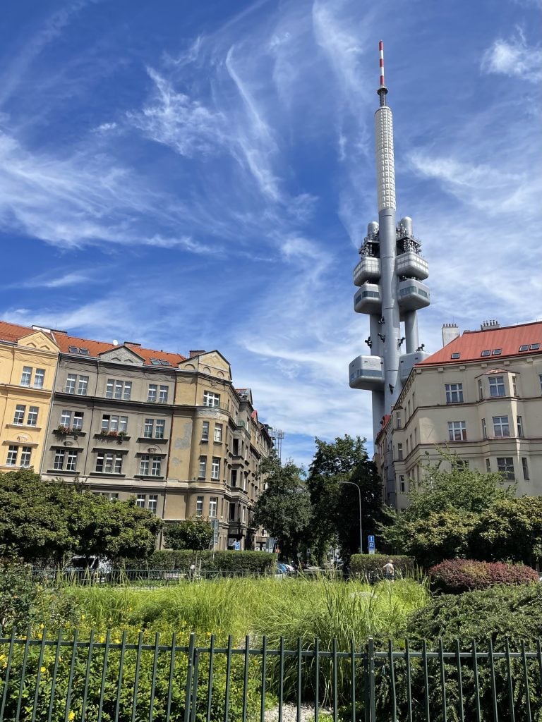 A square with a garden in Prague. You can see warm-colored residential apartments and behind them, the futuristic silver blocky TV tower.