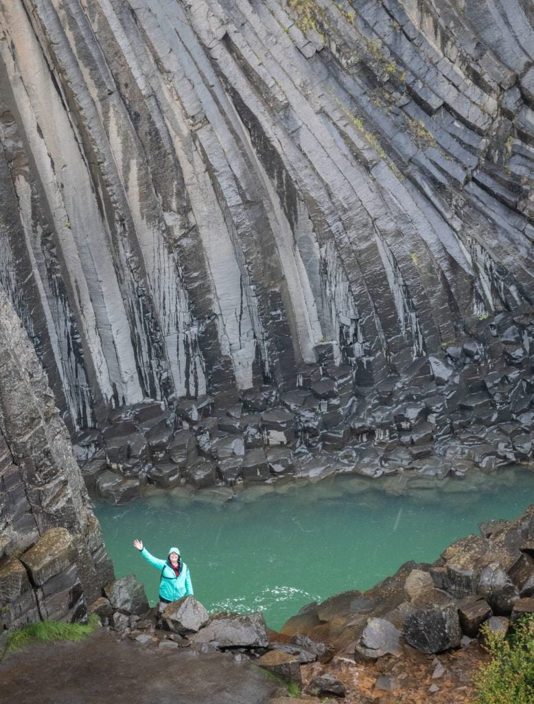 Amanda standing in the rain at the bottom of the canyon near the water, waving and wearing a bright turquoise coat.