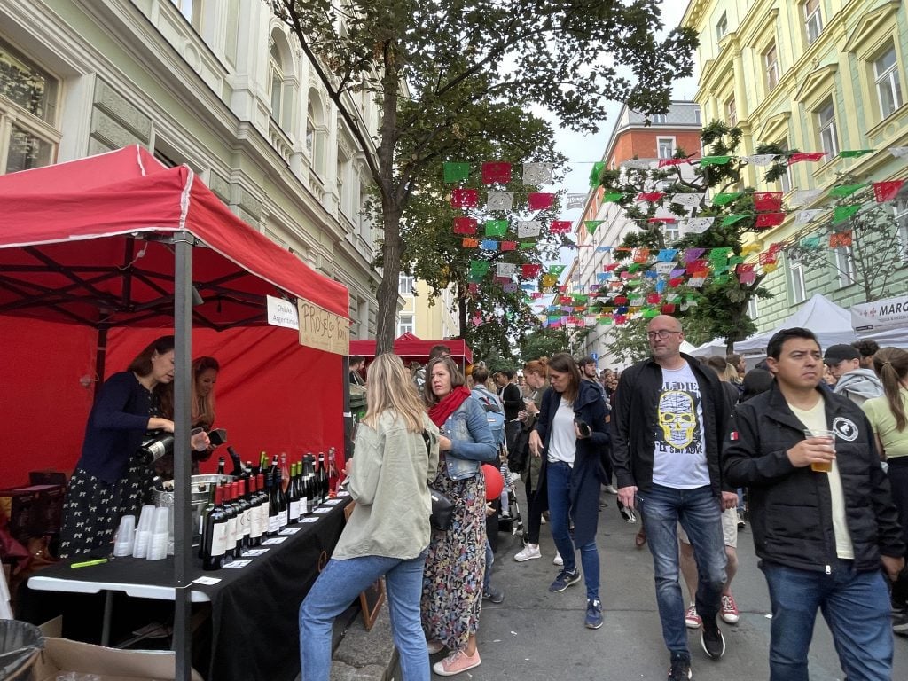 Crowds of people walking down a street in Prague topped with Mexican paper flags, and a street kiosk serving wine.