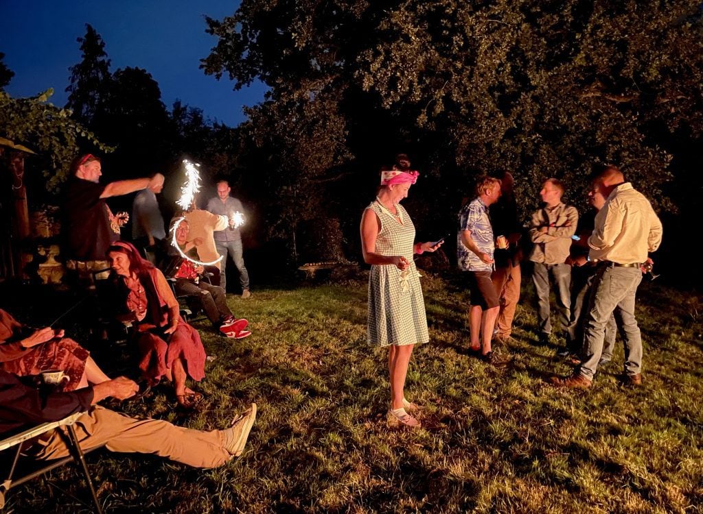 Several people standing around a bonfire in the dark, including one girl in a 50s dress and head wrap, some swirling around sparklers.
