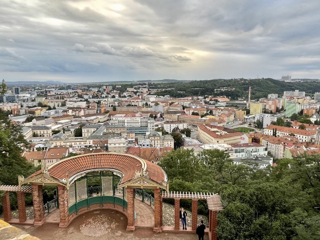 An aerial view over the city of Brno with lots of pastel-colored buildings, most with orange terra cotta roofs, and parks on hills.