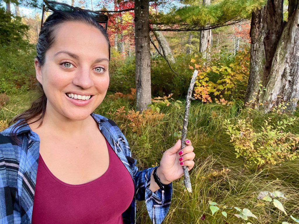 Kate wearing a blue plaid shirt over a burgundy tank top and smiling, holding up a stick in front of lots of vibrant fall foliage.