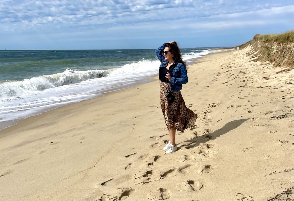 Kate stands on a sandy beach in Martha's Vineyard. She wears a long leopard print skirt, denim jacket, and sunglasses and holds her hair back while looking at the large waves.