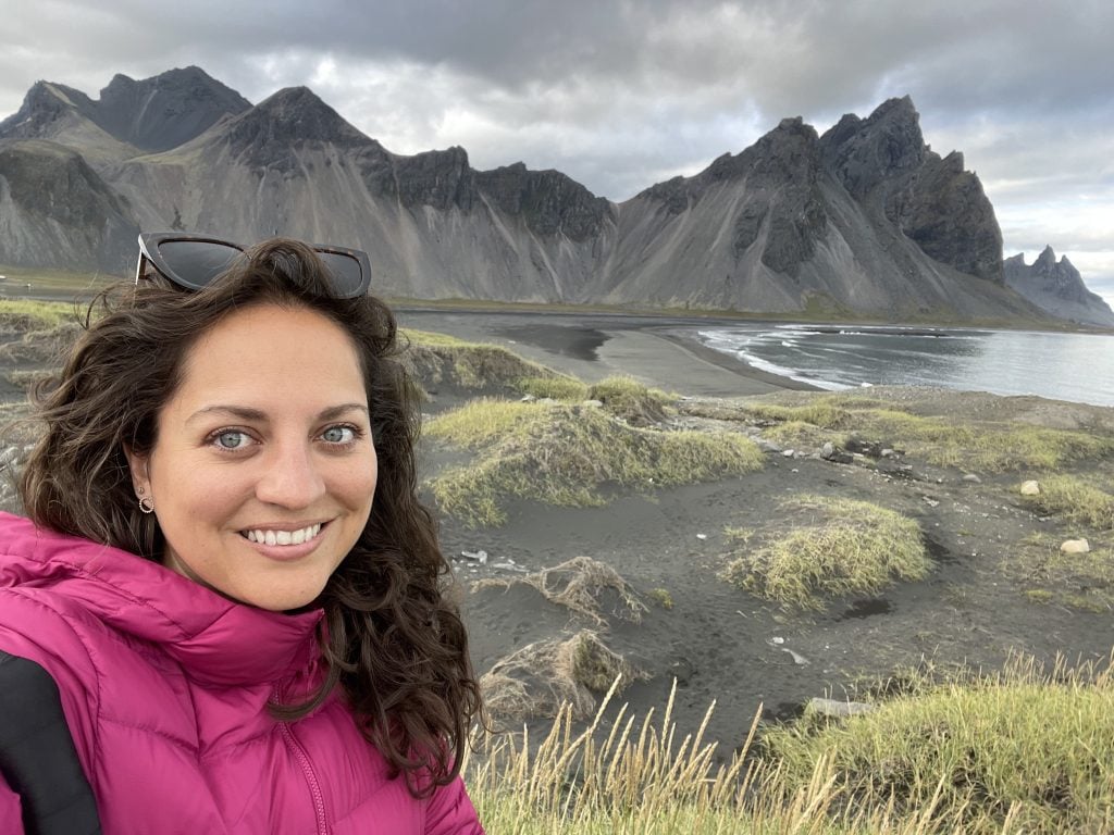 Kate in a pink jacket taking a selfie in front of a black beach studded with grass and jagged black mountains of Stokksnes.