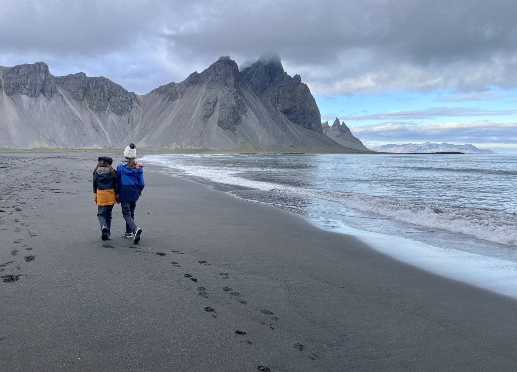 Two children in winter coats walking along the edge of the water, the jagged mountain behind them.