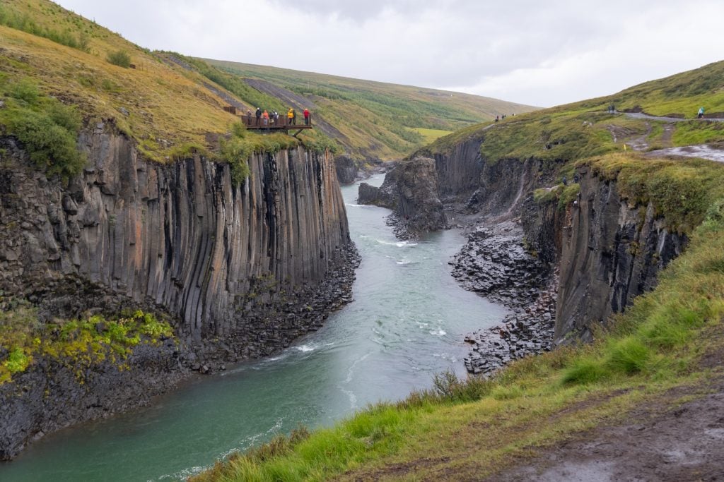 A canyon lined with gray columns of rock, with a turquoise river flowing through it. The sides are grassy and there's a viewing platform on the left.