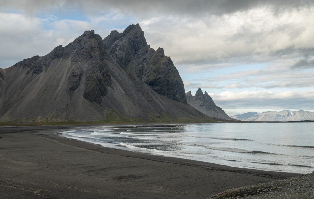 A black beach next to calm water with a black jagged mountain in the background.
