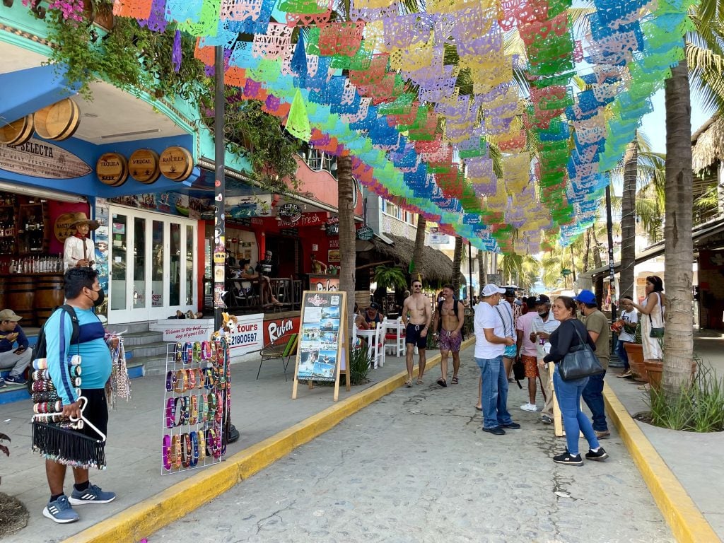 A narrow street lined with stores and street vendors, colorful flags hanging between two rows of palm trees above.