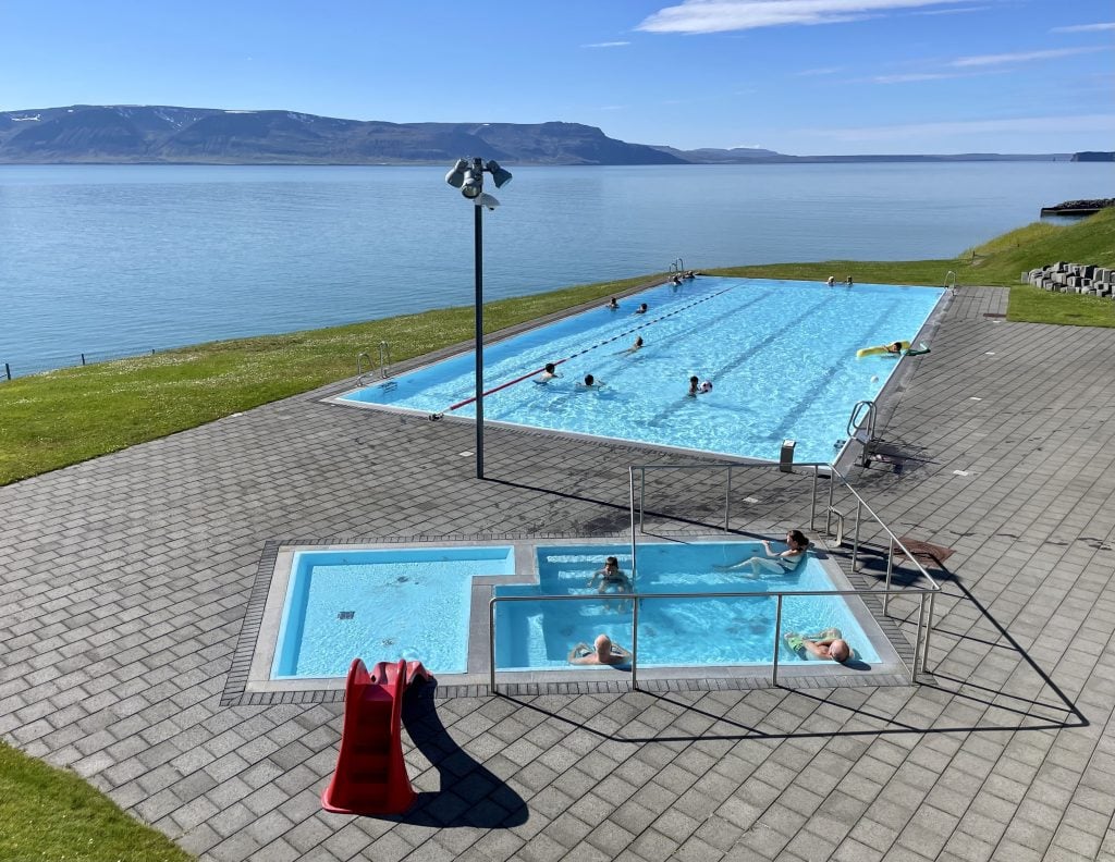 A large swimming pool and a small hot tub, on a grassy hill overlooking a calm blue fjord and mountains in the background.
